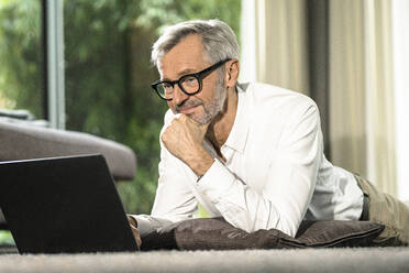 Lächelnder älterer Mann mit grauen Haaren in modernem Design-Wohnzimmer bei der Arbeit am Laptop - SBOF02121