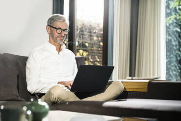 Älterer Mann mit grauen Haaren in modernem Design-Wohnzimmer arbeitet am Laptop im Home-Office - SBOF02123