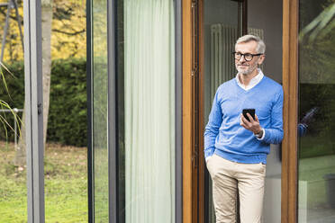 Senior man with grey hair standing in front of his modern design home holding smartphone - SBOF02124