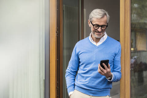 Lächelnder älterer Mann mit grauen Haaren steht vor seinem modernen Designhaus und schaut auf sein Smartphone, lizenzfreies Stockfoto