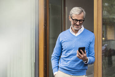 Smiling senior man with grey hair standing in front of his modern design home looking at smartphone - SBOF02125