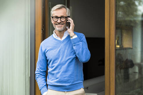 Lächelnder älterer Mann mit grauen Haaren, der vor seinem modernen Designhaus steht und telefoniert - SBOF02126
