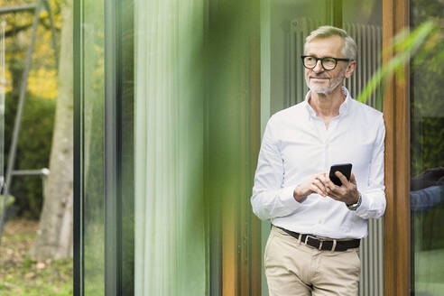 Smiling senior man with grey hair standing in front of his modern design home holding smartphone - SBOF02134