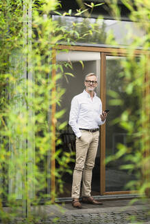 Älterer Mann mit grauem Haar hält Smartphone vor seinem modernen Designhaus im Bambusgarten - SBOF02135
