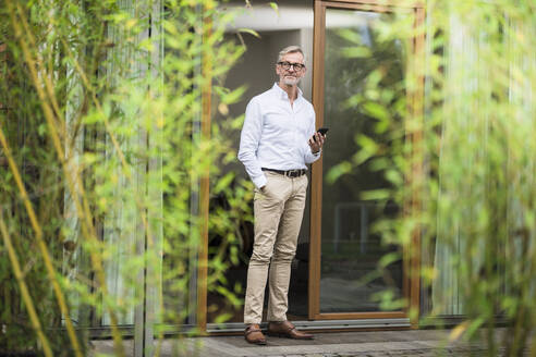 Älterer Mann mit grauen Haaren steht vor seinem modernen Designhaus und hält ein Smartphone in der Hand - SBOF02136