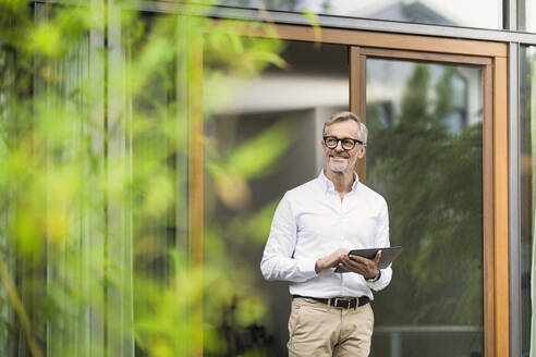 Smiling senior man with grey hair holding tablet in front of his modern design home in bamboo garden - SBOF02137