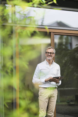 Lächelnder älterer Mann mit grauem Haar, der ein Tablet vor seinem modernen Designhaus im Bambusgarten hält, lizenzfreies Stockfoto