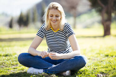 Blond smiling woman sitting on a meadow in a park - JSMF01435