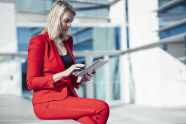 Blonde Geschäftsfrau im roten Anzug und mit digitalem Tablet in einem Bürogebäude - JSMF01441