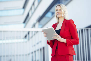 Blond businesswoman wearing red suit and using digital tablet at an office building - JSMF01442