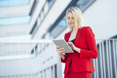 Blonde Geschäftsfrau im roten Anzug und mit digitalem Tablet in einem Bürogebäude - JSMF01443