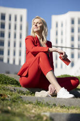 Blond businesswoman wearing red suit and sitting on stairs - JSMF01453