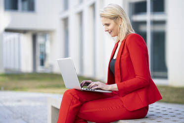 Blond businesswoman wearing red suit and using laptop on a bench - JSMF01455