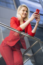 Blonde Geschäftsfrau im roten Anzug, die ihr Smartphone überprüft - JSMF01458
