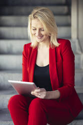 Blonde Geschäftsfrau im roten Anzug und mit digitalem Tablet in einem Bürogebäude - JSMF01462