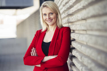 Blond smiling businesswoman with arms crossed wearing red suit and looking at camera - JSMF01463