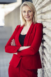 Blond smiling businesswoman with arms crossed wearing red suit and looking at camera - JSMF01464