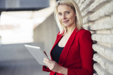 Blond businesswoman wearing red suit and using digital tablet at an office building - JSMF01465