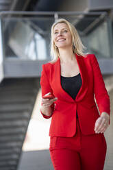 Blond smiling businesswoman wearing red suit and holding smartphone - JSMF01466