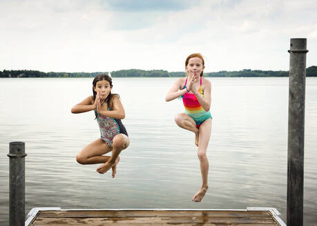 Zwei junge Mädchen in Badeanzügen, die von einem Dock in einen See springen - CAVF75468