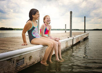 Zwei junge Mädchen in Badeanzügen sitzen lachend auf einem Dock am See - CAVF75469