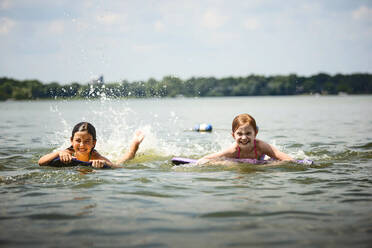Zwei junge Mädchen in Badeanzügen auf Kickboards in einem See - CAVF75471