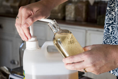 Close up of person standing in a kitchen, decanting liquid from plastic container into glass bottle. - MINF13875