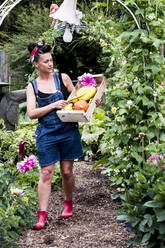 Frau geht in einem Garten spazieren und trägt eine Holzkiste mit frischem Gemüse und Blumen. - MINF13949