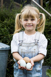 Blondes Mädchen steht in einem Garten, hält frische Eier und lächelt in die Kamera. - MINF13952