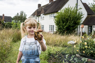 Blondes Mädchen steht in einem Garten und hält eine knubbelige Kartoffel. - MINF13959