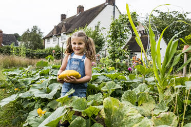 Mädchen steht in einem Gemüsebeet im Garten, hält einen gelben Kürbis und lächelt in die Kamera. - MINF13961