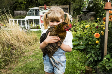 Blondes Mädchen steht in einem Garten und hält ein braunes Huhn. - MINF13966