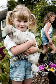 Blondes Mädchen steht in einem Garten und hält ein flauschiges graues Huhn. - MINF13968