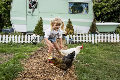 Blondes Mädchen mit zwei Hühnern auf einem Gartenweg, weiß-grüner Retro-Wohnwagen im Hintergrund. - MINF13972