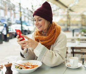 Smiling young woman with smart phone eating lunch at autumn sidewalk cafe - HOXF05036