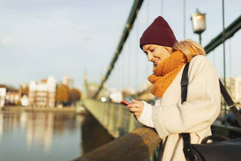 Junge Frau mit Strumpfmütze und Schal benutzt ihr Smartphone auf einer Herbstbrücke - HOXF05041