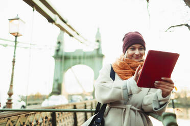 Junge Frau mit Strumpfmütze und Schal benutzt ein digitales Tablet auf einer Brücke in der Stadt - HOXF05057