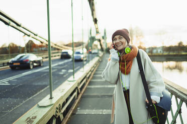 Junge Frau mit Strumpfmütze und Schal spricht auf einer Brücke mit ihrem Smartphone - HOXF05059