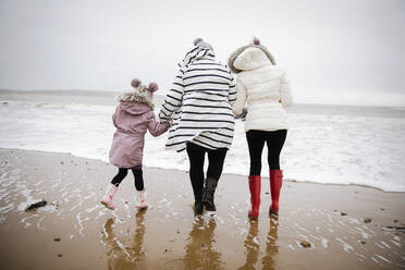 Mother and daughters in warm clothing walking on winter ocean beach - HOXF05091