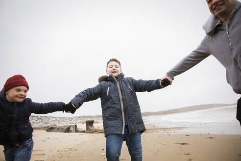 Verspielter Vater und Söhne halten sich am winterlichen Meeresstrand an den Händen - HOXF05100