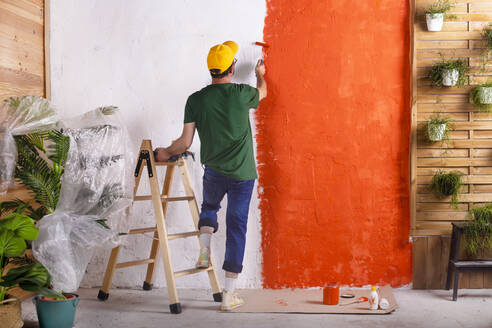 Rear view of man painting orange wall in his garden terrace - RTBF01425