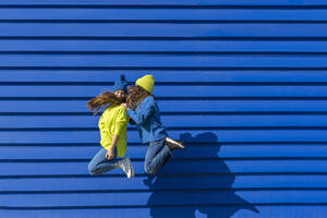 Two teenage girl wearing matching clothes jumping in the air in front of blue background - ERRF02679