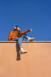 Teenage girl sitting on wall against blue sky listening music with headphones - ERRF02685