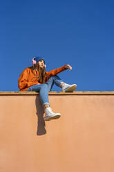 Teenager-Mädchen sitzt an der Wand vor blauem Himmel und hört Musik mit Kopfhörern - ERRF02685