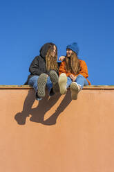Zwei lächelnde Teenager-Mädchen sitzen an einer Wand vor blauem Himmel - ERRF02686