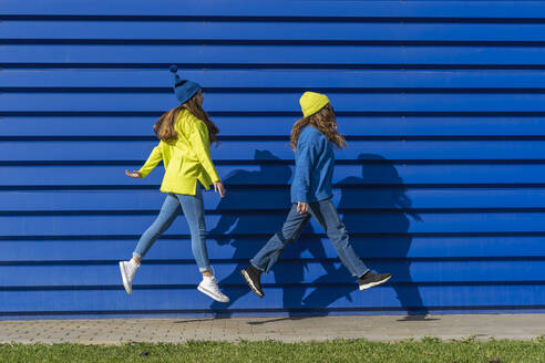 Two teenage girls wearing matching clothes jumping in the air in front of blue background - ERRF02693