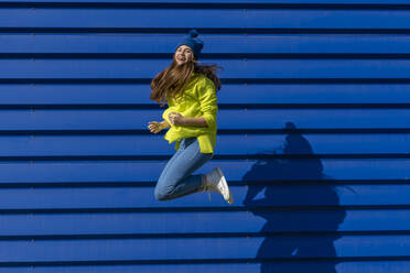 Lachendes Teenager-Mädchen, das vor einem blauen Hintergrund in die Luft springt - ERRF02696