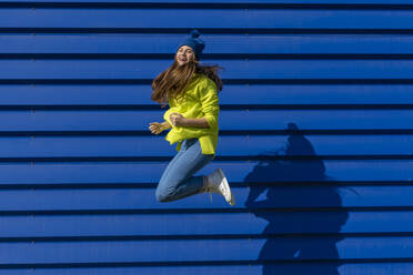 Laughing teenage girl jumping in the air in front of blue background - ERRF02696