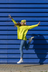 Happy teenage girl jumping in the air in front of blue background listening music with headphones - ERRF02698