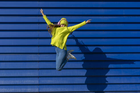 Teenager-Mädchen, das vor einem blauen Hintergrund in die Luft springt und mit Kopfhörern Musik hört - ERRF02701